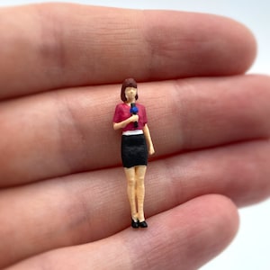 Pode incluir: Uma figura em miniatura de uma pessoa segurando um microfone, vestindo uma camisa vermelha, uma saia preta e sapatos pretos.