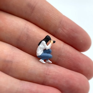Pode incluir: Uma figura em miniatura de uma pessoa agachada e segurando uma c&acirc;mera. A pessoa est&aacute; vestindo uma camisa branca, jeans azul e cabelo preto.