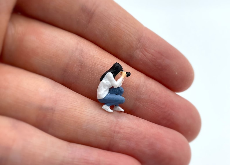 Pode incluir: Uma figura em miniatura de uma pessoa agachada e segurando uma c&acirc;mera. A pessoa est&aacute; vestindo uma camisa branca, jeans azul e cabelo preto.