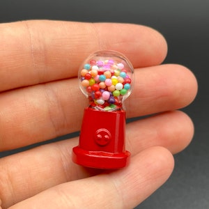 Op de afbeelding: Een miniatuur rode kauwgomballenautomaat met een doorzichtige, afgeronde bovenkant gevuld met kleurrijke kauwgomballen. De basis van de machine heeft een klein, lachend gezichtsontwerp. De kauwgomballen zijn in verschillende kleuren, waaronder rood, geel, blauw en groen.