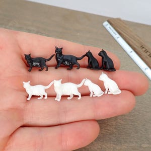 Może przedstawiać: Miniaturowe figurki czarnych i białych kotów. Figurki są w różnych pozach, w tym stojących i siedzących. Drewniana linijka znajduje się w tle.