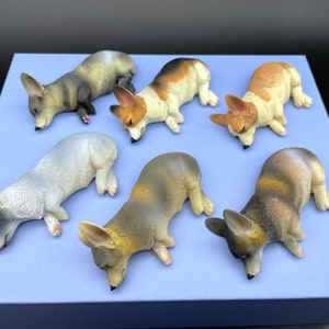 Puede incluir: Seis figuritas de corgi durmiendo en varios colores, incluyendo gris, marrón, blanco y naranja, sobre un fondo azul.