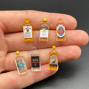Op de afbeelding: Zes miniatuur flessen sterke drank met verschillende etiketten, waaronder Jim Beam, Black Velvet, Famous Grouse en Torys. De flessen worden in een hand gehouden.