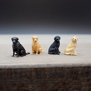 Puede incluir: Cuatro figuritas de perros en miniatura, dos labradores negros y dos labradores amarillos. Los perros están sentados y mirando hacia adelante.