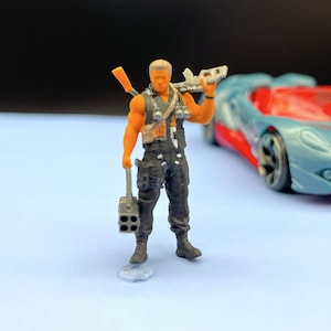 Può includere: Una piccola action figure dettagliata di un soldato che indossa un giubbotto nero e tiene una pistola e un lanciarazzi. La figura ha un colorito abbronzato e capelli castano corti. La figura è in piedi su una superficie blu.