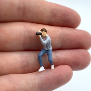 Pode incluir: Uma figura em miniatura de uma pessoa vestindo um su&eacute;ter cinza e jeans azul, segurando uma c&acirc;mera e olhando atrav&eacute;s do visor.