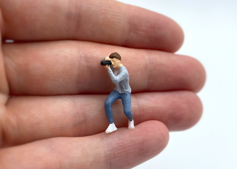 Pode incluir: Uma figura em miniatura de uma pessoa vestindo um su&eacute;ter cinza e jeans azul, segurando uma c&acirc;mera e olhando atrav&eacute;s do visor.