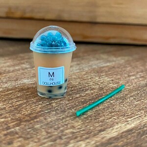 May include: A miniature plastic cup filled with a brown liquid and blue boba pearls. The cup has a white label that says "M ini DOLLHOUSE". A green plastic straw is on the wooden surface.