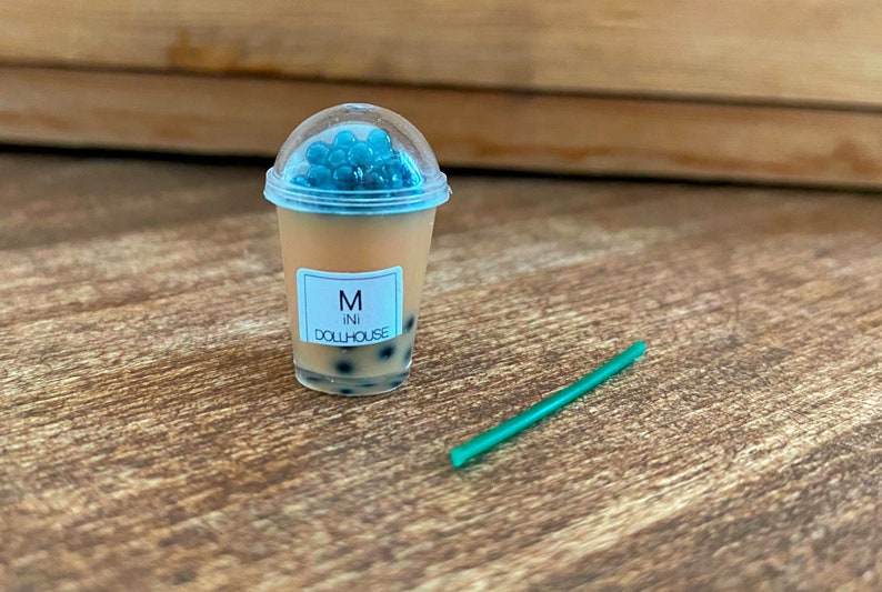 May include: A miniature plastic cup filled with a brown liquid and blue boba pearls. The cup has a white label that says "M ini DOLLHOUSE". A green plastic straw is on the wooden surface.