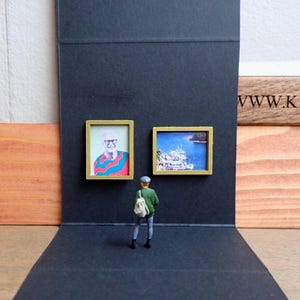 Puede incluir: Escena de galería de arte en miniatura con dos pinturas enmarcadas en una pared negra. Una pequeña figura con chaqueta y sombrero verdes se encuentra frente a las obras de arte. Las pinturas representan un retrato y un paisaje marino. Paneles de madera enmarcan la escena.
