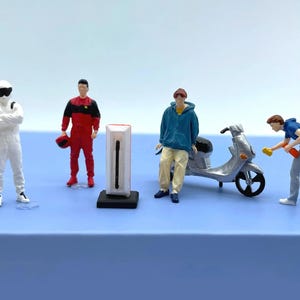 Puede incluir: Figuras en miniatura de personas con diferentes atuendos, incluyendo un traje de carreras y una sudadera con capucha azul, dispuestas alrededor de un scooter plateado y un modelo de estación de carga. La escena se desarrolla sobre una superficie azul.