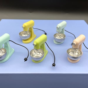 May include: Five miniature stand mixer kitchen appliances in different colours: yellow, green, light blue, pink, and blue. Each mixer has a silver bowl and a silver whisk attachment.