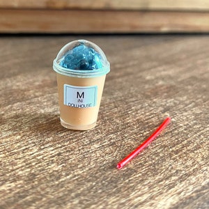 May include: A miniature plastic cup with a clear lid and a brown liquid inside. The cup has a white label with the text "M ini DOLLHOUSE". A red plastic straw is lying on a wooden surface.