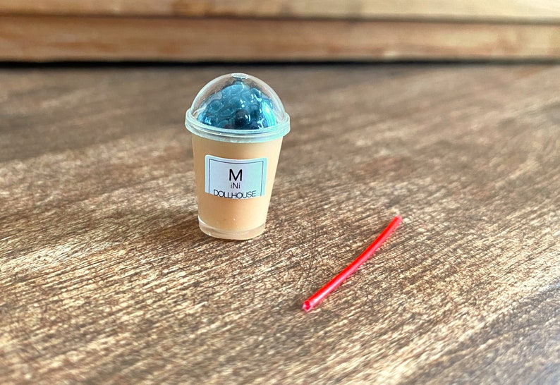 May include: A miniature plastic cup with a clear lid and a brown liquid inside. The cup has a white label with the text "M ini DOLLHOUSE". A red plastic straw is lying on a wooden surface.