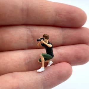 Pode incluir: Uma figura em miniatura de uma pessoa agachada segurando uma c&acirc;mera. A figura &eacute; feita de pl&aacute;stico e &eacute; pintada em cores realistas. A pessoa est&aacute; vestindo uma camisa preta e shorts verdes. A c&acirc;mera &eacute; preta e tem uma lente.