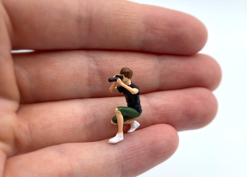Pode incluir: Uma figura em miniatura de uma pessoa agachada segurando uma c&acirc;mera. A figura &eacute; feita de pl&aacute;stico e &eacute; pintada em cores realistas. A pessoa est&aacute; vestindo uma camisa preta e shorts verdes. A c&acirc;mera &eacute; preta e tem uma lente.