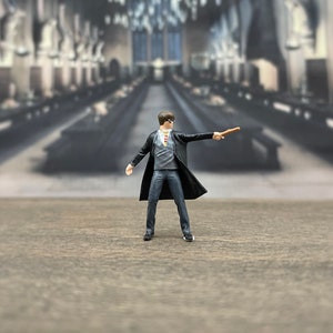 May include: A figurine of a young wizard wearing a black robe and glasses, holding a wand in his right hand. The figurine is standing on a wooden surface.