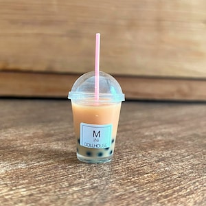 May include: A miniature plastic cup filled with a brown liquid and black boba pearls. The cup has a clear lid and a pink straw. The cup has a white label that says "Mini Dollhouse" and "M" in a circle.