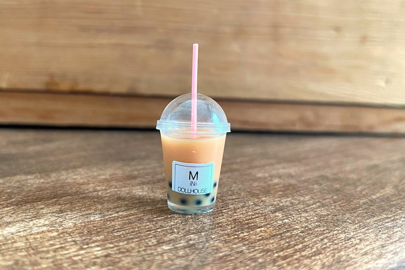May include: A miniature plastic cup filled with a brown liquid and black boba pearls. The cup has a clear lid and a pink straw. The cup has a white label that says "Mini Dollhouse" and "M" in a circle.