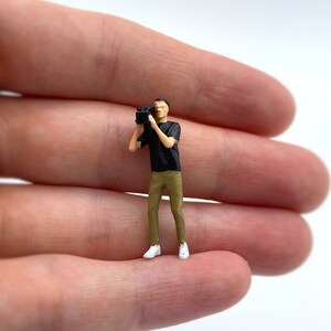 Pode incluir: Uma figura em miniatura de uma pessoa segurando uma c&acirc;mera, vestindo uma camisa preta e cal&ccedil;as c&aacute;qui. A figura est&aacute; em p&eacute; sobre uma m&atilde;o.