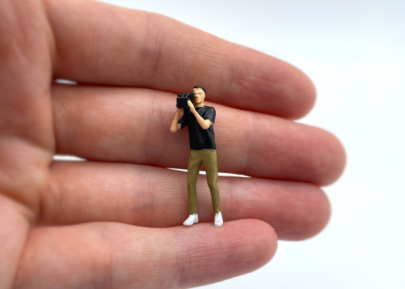 Pode incluir: Uma figura em miniatura de uma pessoa segurando uma c&acirc;mera, vestindo uma camisa preta e cal&ccedil;as c&aacute;qui. A figura est&aacute; em p&eacute; sobre uma m&atilde;o.