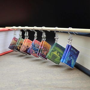 Peut inclure: Une série de sept boucles d'oreilles miniatures en forme de livre, chacune avec une couverture différente, suspendues à des crochets argentés. Les boucles d'oreilles sont présentées sur fond de livres et de fond sombre.