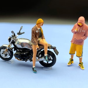 Könnte beinhalten: Ein silbernes Motorrad mit einer weiblichen Figur, die auf dem Sitz sitzt. Eine männliche Figur in einem rosa Hoodie und gelben Shorts steht rechts neben dem Motorrad.