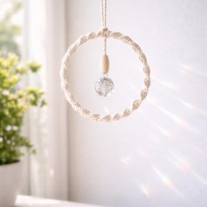 Könnte beinhalten: Ein Makramee-Kristallfänger mit einem klaren, facettierten Kristall und einer Holzperle. Das kreisförmige Design besteht aus cremefarbenem, gedrehtem Baumwollseil. Der Sonnenfänger hängt an einer cremefarbenen Kordel und erzeugt eine Boho-Ästhetik.