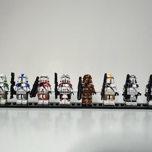 May include: A set of eight plastic toy soldier minifigures standing on a black rectangular base. Each figure wears a white helmet with distinct colored markings including green, blue, red, maroon, brown, yellow, dark blue, and purple, holding black blasters.