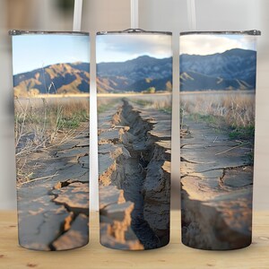 May include: Three stainless steel tumblers with a design of a cracked dirt road leading to a mountain range in the distance. The road is cracked and dry, with a hint of green grass on the sides.