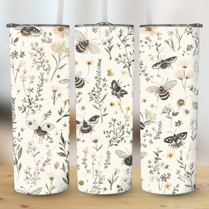 Op de afbeelding: Drie roestvrijstalen bekers met een witte achtergrond en een patroon van bijen en bloemen. De bekers zijn allemaal even groot en hebben een deksel.
