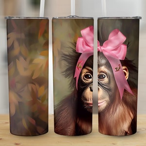 Puede incluir: Tres vasos de acero inoxidable con diferentes diseños. El primer vaso tiene un patrón abstracto de hojas marrones y naranjas. El segundo y tercer vaso presentan un primer plano de un orangután con un lazo rosa. El orangután tiene pelaje marrón y una expresión seria.