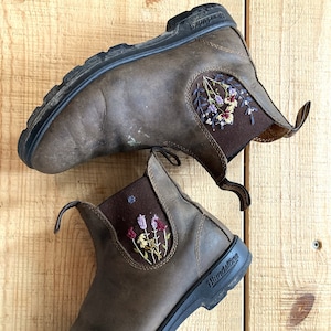 Two Blundstone Chelsea boots laying on  a wooden backdrop. Each boot is adorned with custom floral embroidery on the  outer elastic panel.