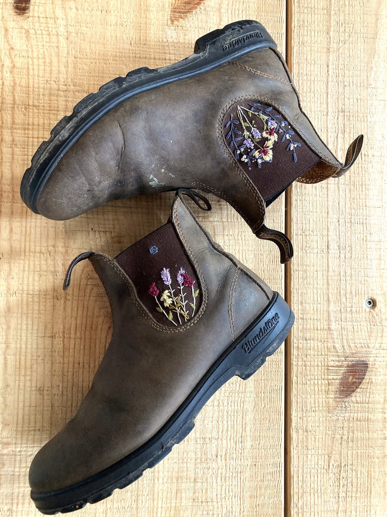 Two Blundstone Chelsea boots laying on  a wooden backdrop. Each boot is adorned with custom floral embroidery on the  outer elastic panel.