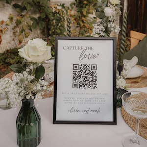 Könnte beinhalten: Ein schwarzer gerahmter Schild mit dem Text "CAPTURE THE love" und einem QR-Code. Das Schild steht auf einem Tisch, der für eine Feier gedeckt ist, mit Blumen, Kerzen und Glaswaren. Das Schild enthält auch die Namen "olivia und noah".