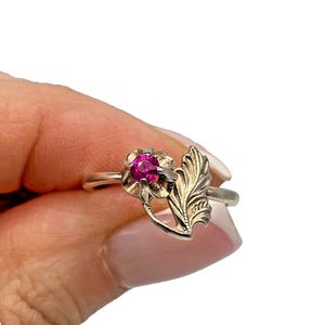 May include: A silver ring with a flower-shaped setting holding a bright pink gemstone. An ornate leaf design is on the side. The ring's polished finish and intricate details create an elegant aesthetic. A piece of jewellery.