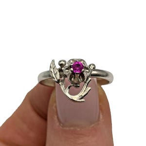 Op de afbeelding: Een zilveren ring met een bloemmotief en een helder roze edelsteen. De ring heeft een gedetailleerd bloemmotief met bladeren en knoppen. De edelsteen heeft een levendige roze kleur, gezet in het midden van de bloem.