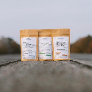 May include: Three tan coffee bags with airplane illustrations and text labels. The bags are labeled "ON THE STEP", "SURFIN' CLOUDS", and "THUNDERBOLT". Each bag has a white label with additional text and color-coded bars indicating roast level and weight.