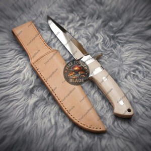 Könnte beinhalten: Ein feststehendes Messer mit einer polierten Stahlklinge, einem hellen Griff mit zwei silbernen Nieten und einer beigen Lederscheide. Ein kreisförmiges Logo mit einem Amboss und dem Text Fire Forged Blade ist mittig auf der Klinge platziert.