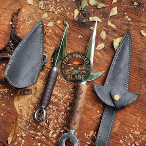 Könnte beinhalten: Zwei Wurfmesser aus Metall mit spitzen Klingen und Ringknäufen. Ein Messer hat einen dunkelbraun umwickelten Griff, das andere einen hellbraunen Holzgriff mit Gravuren. Jedes Messer enthält eine schwarze, strukturierte Scheide mit gelben Nähten.