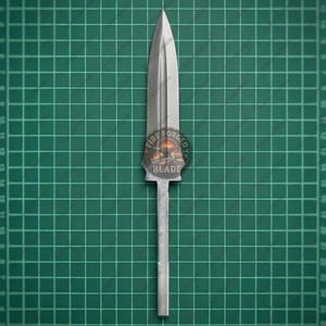 Puede incluir: Una punta de lanza de metal plateado con un extremo afilado y una cresta central a lo largo de la hoja. La base presenta una espiga rectangular alargada para su fijación. Un logotipo circular con el texto FIRE FORGED BLADE es visible.