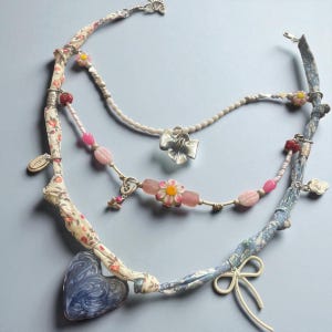 May include: A multi-strand necklace featuring fabric-wrapped strands with floral patterns, adorned with beads, charms, and a heart-shaped pendant. The necklace includes a silver bow pendant and a delicate flower design.