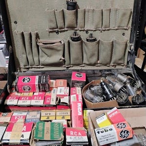 May include: Open black vintage suitcase filled with various electronic tubes and components. The case has a green fabric interior with pockets. Boxes of RCA and Sylvania tubes are visible, along with loose tubes in a small basket.