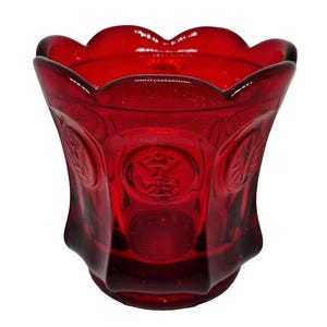 May include: A vibrant red glass votive holder featuring a scalloped rim and a series of embossed circular medallions around the exterior. The glass has a translucent quality, and the molded relief patterns depict various heraldic-style symbols.