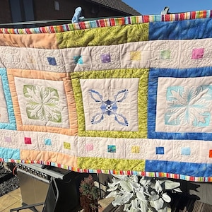 May include: A colourful patchwork quilt, featuring embroidered floral designs within square blocks. The quilt's palette includes blue, green, orange, and cream, with a striped and solid-coloured border. The quilt is displayed outdoors, showcasing its handmade quality and vibrant colours.