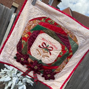 May include: A festive Christmas wall hanging with a cream background and a red border. The centre features a square with "Merry Christmas" and bells embroidered. Burgundy fabric flowers and plaid ribbon add detail.