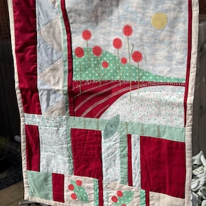 May include: A handmade fabric wall hanging with a landscape design. The quilt has a red, green, and cream colour scheme with appliqued red flower details, a yellow sun, and a green dotted hill. The quilt is hanging outside.
