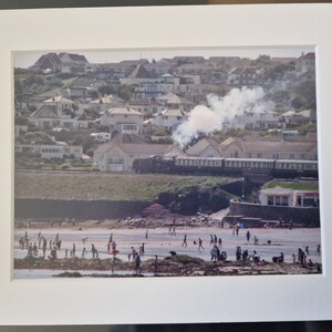 May include: A framed photograph of a beach scene with a steam train passing along the coast. People are on the beach, and houses are on the hillside in the background. The photograph is in a white mat.