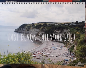 Könnte beinhalten: Ein Kalender für 2027 mit einem Foto der Küste von East Devon. Das Bild zeigt einen Strand mit Booten, Klippen und einem bewölkten Himmel. Der Text "EAST DEVON CALENDAR 2027" ist in Weiß gedruckt.