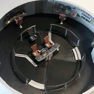 Puede incluir: Un modelo del interior del puente de una nave espacial con un suelo gris y negro, un panel de control y varias sillas. El puente está rodeado por una pared blanca.